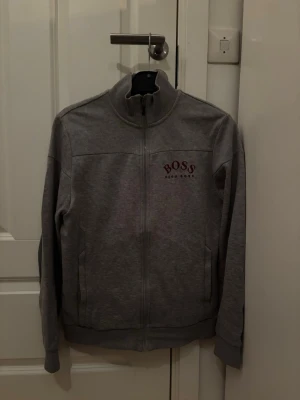 Grå ziptröja från Hugo Boss - Snygg grå ziptröja från Hugo Boss med hög krage och diskret logga i vinrött på bröstet. Tröjan har två fickor framtill och ribbade muddar vid ärmslut och nederkant. Perfekt för en avslappnad och stilren look.