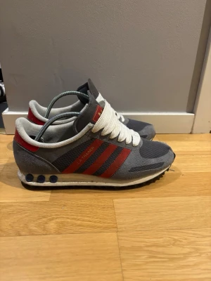 Adidas LA Trainer grå/röd sneakers - Snygga Adidas LA Trainer sneakers i grått med röda detaljer och klassiska tre ränder på sidan. Ovandelen är i mesh och mocka, med vita skosnören och retrovibbar. Sulan har de ikoniska pluggarna på sidan. Perfekta för dig som gillar streetstyle och sportig look.