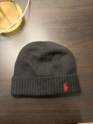 Svart mössa från Polo Ralph Lauren - Klassisk svart ribbstickad mössa från Polo Ralph Lauren med uppvikt kant och röd broderad logga framtill. Tillverkad i mjukt stickat material som håller dig varm och snygg. Perfekt accessoar för kyliga dagar.