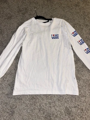 Vit långärmad Vans t-shirt - Vit långärmad t-shirt från Vans med trycket 'I ❤️ MY VANS' i blått och rött på bröstet och längs ena ärmen. Klassisk passform och rund halsringning. Perfekt för dig som gillar streetwear och vill ha en clean look med tydlig logga.