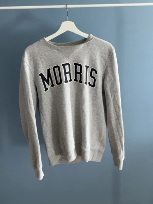 Grå sweatshirt från Morris - Snygg grå sweatshirt från Morris med stor svartvit logga på bröstet. Tröjan har rund halsringning, ribbade muddar vid ärmslut och nederkant samt en klassisk college-look. Perfekt för en avslappnad och trendig stil.