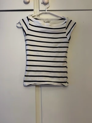 Randig vit och svart topp - Snygg vit topp med svarta horisontella ränder och korta ärmar. Modellen är figurnära och har en klassisk, enkel design som passar till mycket. Perfekt för dig som gillar en stilren och tidlös look.