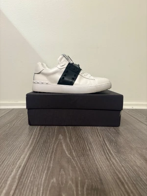 Vita Valentino sneakers med nitar - Snygga vita sneakers från Valentino med svarta detaljer och coola nitar på sidan. Skorna har en låg profil och är tillverkade i läder med en chunky vit sula. Perfekta för dig som vill sticka ut med en exklusiv och trendig look. Replica