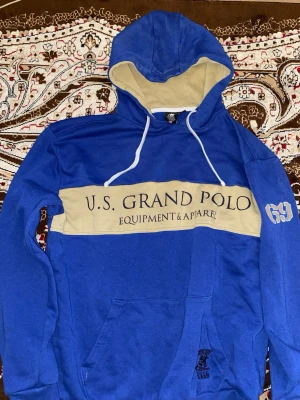 Blå hoodie från U.S. Grand Polo - Blå hoodie med beige detaljer och stor huva från U.S. Grand Polo Equipment & Apparel. Stor beige panel över bröstet med tryckt logga och text, samt siffran 69 på ärmen. Känguruficka framtill och vita dragsnören i huvan. Skön och avslappnad stil.