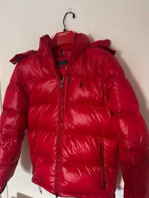 Röd pufferjacka från Polo Ralph Lauren - Snygg röd pufferjacka från Polo Ralph Lauren med blank finish, huva och dragkedjor framtill. Jackan har en klassisk pufferdesign med broderad logga på bröstet och flera praktiska fickor. Perfekt för kalla dagar när du vill hålla stilen.