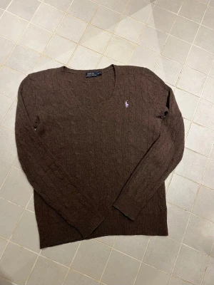 Brun kabelstickad tröja Polo Ralph Lauren - Mysig brun kabelstickad tröja från Polo Ralph Lauren med v-ringning och lång ärm. Tröjan har ett litet broderat logotypmärke i lila på bröstet och ribbade muddar vid ärmslut och nederkant. Perfekt för lager på lager under kyliga dagar.