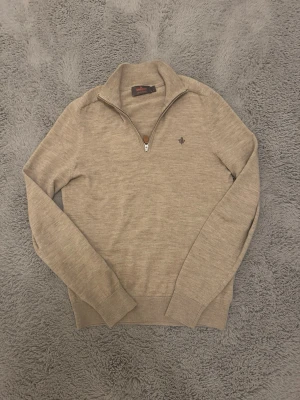 Beige halfzip tröja från Morris - Snygg beige stickad tröja från Morris med halv dragkedja och broderad lilja på bröstet. Tröjan har lång ärm, ribbade muddar och är tillverkad i mjukt material som känns skönt mot huden. Perfekt för lager på lager och stilrena outfits.