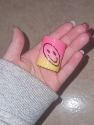 Fidget leksak - Säljer en liten plastfjäder i rosa och gult med en svart smiley på. Perfekt som leksak eller dekoration. Fjädern är flexibel och kan dras ut och tryckas ihop, hoppar ner på trappstegen. Enkel att ta med och rolig att använda.