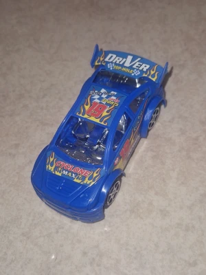 Leksaksbil - Säljer en blå leksaksbil i plast med racingtema. Bilen har dekaler med texten 'Cyclone Max', 'Driver Top-Hole' och siffran 19, samt gula eldflammor på huven och spoilern. Sportig design med silverfärgade detaljer och svarta hjul.