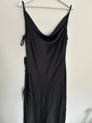 Svart balklänning från ASOS DESIGN - Elegant svart balklänning i satin med slit och tre dekorativa rosetter längs ena sidan som ger en exklusiv och feminin detalj ✨  Perfekt till bal, gala, bröllop eller andra festliga tillfällen 🖤  Klänningen har ett vackert fall och känns lyxig i materialet. Klänningen är helt ny och orginalförpackning medföljer.  Jag är 178 cm lång och bär vanligtvis M och den går nästan ned till fotknölarna, perfekt längd för klackar 👠 