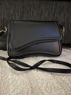 Svart axelväska med vågig kant - Snygg svart axelväska i konstläder med unik vågig kant på locket och guldfärgade detaljer. Väskan har magnetlås och en lång axelrem som kan bäras crossbody. Perfekt för dig som gillar stilrena accessoarer med en twist.