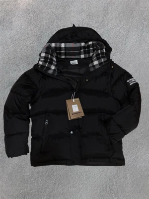 Svart pufferjacka från Burberry - Snygg svart pufferjacka från Burberry med huva och klassiskt rutigt foder i svart, vitt och rött. Jackan är storlek L men passar som M. Den har dragkedja, tryckknappar och två fickor framtill. Burberry-logga på ärmen och justerbar huva. Perfekt för kalla vinterdagar eller fester.