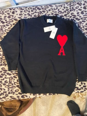 Svart stickad tröja från Ami - Säljer en svart stickad tröja från Ami med deras ikoniska röda hjärta och bokstaven A broderat på bröstet. Tröjan har rund hals, ribbade muddar och är i mjukt material. Perfekt för dig som gillar stilrena plagg med en twist.