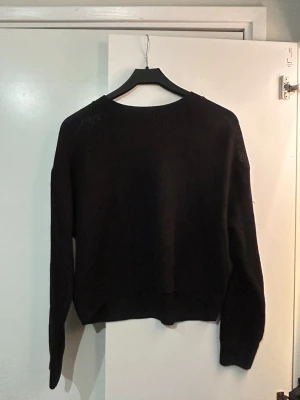 Svart stickad tröja med rund hals - En enkel svart stickad tröja med rund hals och långa ärmar. Tröjan har en rak passform och ribbade muddar vid ärmslut och nederkant. Tröjan är åt det tunnare hållet!