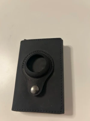 Svart plånbok i läder - Säljer en svart minimalistisk plånbok i läder med en rund hållare på framsidan, perfekt för AirTag eller liknande tillbehör. Plånboken har en stilren och kompakt design som gör den enkel att bära med sig i fickan eller väskan.