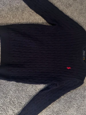 Mörkblå kabelstickad tröja Polo Ralph Lauren - Snygg mörkblå kabelstickad tröja från Polo Ralph Lauren med klassisk röd logga broderad på bröstet. Tröjan har rund halsringning och långa ärmar. Perfekt för dig som gillar tidlös och stilren design.