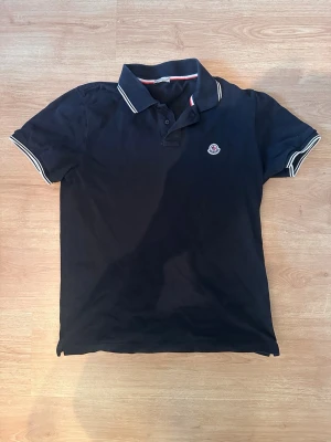 Svart Moncler piké  - Schyst monclizzy pike, skickas samma dag eller dagen efter, Ser lite skrynklig ut eftersom den bara har legat i garderoben, Annars använd max några dagar förra året