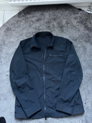 Mörkblå vindjacka från J.Lindeberg - Snygg mörkblå vindjacka från J.Lindeberg med dragkedja framtill, bröstficka med dragkedja och klassisk krage. Tillverkad i lätt och slitstarkt material, perfekt för blåsiga dagar. Diskret logga på bröstet och innerficka med dragkedja för extra förvaring.