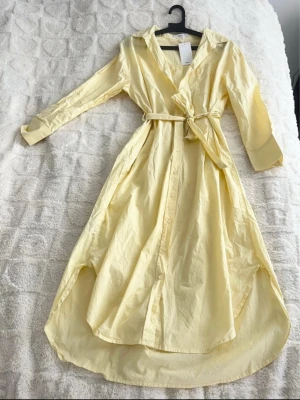 Ljusgul skjortklänning med bälte, bomull XS - Snygg ljusgul bomull midilänning i skjortmodell med knappar framtill och matchande bälte i midjan. Klänningen har lång ärm och klassisk krage, perfekt för en fräsch och stilren look. Tillverkad i mjuk bomull som känns skön mot huden.