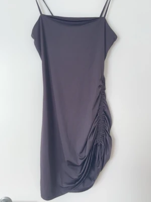 Svart kort klänning med drapering - Säljer en svart kort klänning med tunna axelband och snygg justerbar knytning på sidan. Klänningen har en tight passform och är perfekt för dig som gillar minimalistisk stil med en twist. Materialet känns mjukt och följsamt mot kroppen.