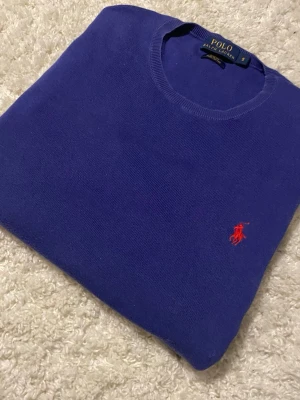 Blå stickad tröja från Polo Ralph Lauren - Säljer en lila stickad tröja från Polo Ralph Lauren i slim fit-modell. Tröjan är gjord av pima cotton och har rund hals samt den klassiska röda logotypen broderad på bröstet. Perfekt för dig som gillar stilrena och tidlösa plagg.