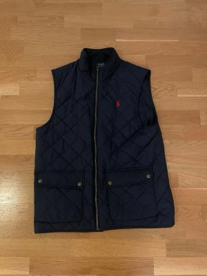 Marinblå dunväst från Polo Ralph Lauren - Snygg marinblå dunväst från Polo Ralph Lauren med quiltat mönster, två stora fickor med lock och tryckknappar samt dragkedja framtill. Klassisk röd logga broderad på bröstet. Perfekt för lager på lager och streetwear-stil.