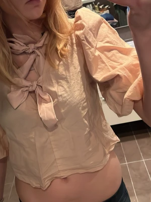 Ljusrosa blus med puffärm och rosetter - Trendig ljusrosa blus med puffiga korta ärmar och två stora rosetter framtill. Blusen har en lös passform och är tillverkad i ett tunt, lätt material som ger en luftig känsla. Perfekt för dig som gillar en söt och unik stil.