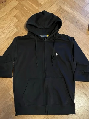 Svart hoodie från Polo Ralph Lauren - Snygg svart hoodie från Polo Ralph Lauren med dragkedja, huva med dragsko och klassisk logga broderad på bröstet. Helt perfekt skick och är så skön att ha på sig, 100% äkta som man kan se på qr koden (bild 3) nypris runt 1999 kr. Priset är inte fastspikat och tvivla inte på att fråga något om du har någon fundering. STORLEK: M/ passar även S
