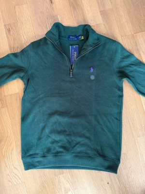 Grön half-zip tröja från Polo Ralph Lauren - Mörkgrön ribbad tröja från Polo Ralph Lauren med halv dragkedja och lila broderad logga på bröstet. Klassisk passform med hög krage och muddar vid ärmslut och nederkant. Perfekt för en stilren och avslappnad look.