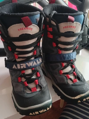 Airwalk Freeride snowboardkängor - Säljer ett par grymma snowboardkängor från Airwalk, modell Freeride. De är svarta och grå med röda detaljer och blått spänne. Kraftig sula och robust design, perfekta för vinterns äventyr. Snygg logga på sidan och framtill. Sköna att ha på sig och riktigt coola.