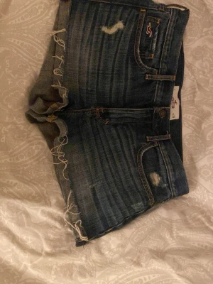 Mörkblå jeansshorts från Hollister - Säljer ett par mörkblå jeansshorts från Hollister med råa kanter och slitningar både fram och bak. Shortsen har klassisk femficksdesign, låg midja och detaljerade sömmar på bakfickorna. Perfekta för sommaren! Köpte här på Plick för 300kr men dom passade inte