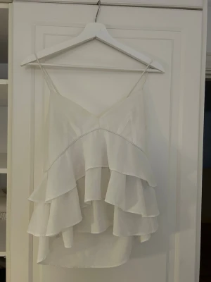 Vit volangig linnetopp - Säljer en vit linnetopp med tunna axelband och flera lager volanger framtill. Toppen har en luftig och flowy känsla, perfekt till sommaren