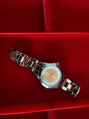 Silverfärgad klocka med roséguld - Stilren klocka med metallarmband i silver och roséguld. Urtavlan är roséguldig med markerade timmar och små stenar runt om. Klockan har en klassisk rund form och metallisk finish, perfekt för dig som gillar en elegant accessoar.