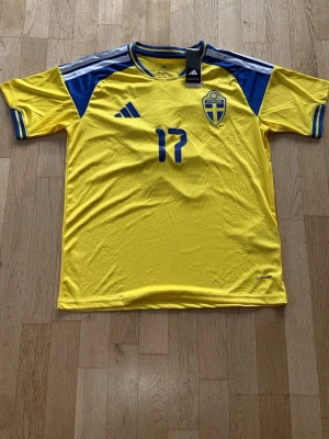 Sverige matchtröja Adidas gul/blå - Säljer en officiell Sverige matchtröja från Adidas i gult och blått med nummer 17 på bröstet. Tröjan har klassiska Adidas-ränder på axlarna, broderat landslagsemblem och rund hals. Tillverkad i lätt och ventilerande material, perfekt för fotboll.
