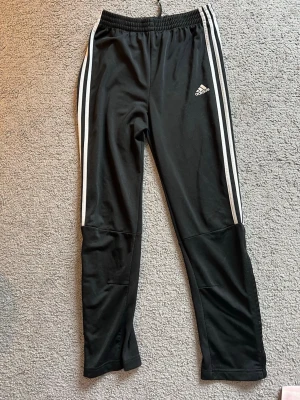 Svarta Adidas träningsbyxor - Säljer ett par svarta träningsbyxor från Adidas med klassiska vita ränder längs sidorna och Adidas-logga på låret. Byxorna har dragkedja vid bensluten och resår i midjan. Storleken är 12-14 år så ca 158-164.