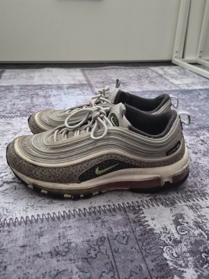 Nike Air Max 97 grå sneakers - Nike Air Max 97 sneakers i grått med reflekterande detaljer och grönt Swoosh-märke på sidan. Ovandelen är i mesh och syntet med vågiga linjer och synlig Air-enhet i sulan. Skorna har snörning och svart innerfoder. Perfekt för dig som gillar streetwear.