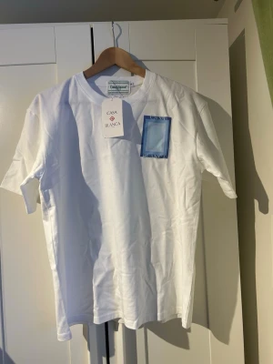 Vit t-shirt från Casablanca med tryck - Säljer en vit t-shirt från Casablanca med blått grafiskt tryck på bröstet och ryggen. T-shirten har rund hals och korta ärmar. Materialet är mjuk bomull och passformen är normal. Perfekt för dig som gillar stilrena plagg med en twist. Helt ny priset kan diskuteras 