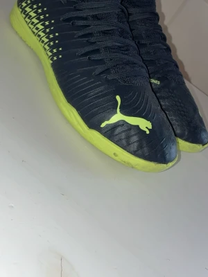 Puma fotbollsskor svart/gul - Säljer ett par snygga fotbollsskor från Puma i svart med neon-gula detaljer och sula. Skorna har snörning och den klassiska Puma-loggan på sidan. Ovandelen är i syntetmaterial och designen är modern och sportig, perfekt för inomhusspel.