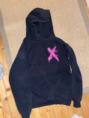 Svart hoodie med rosa tryck - Svart hoodie med huva och magficka. På bröstet finns ett stort rosa X och på ryggen står det 'THE GOOD DIE YOUNG' i rosa graffiti-stil. Hoodien har en avslappnad passform och är tillverkad i mjukt bomullsmaterial. Perfekt för dig som gillar streetwear och statement-plagg.