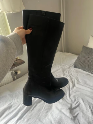 Svarta höga läderstövlar med klack - Snygga svarta stövlar i äkta läder med bred klack och dekorativa broguedetaljer vid tån. Stövlarna har dragkedja på insidan och når upp till knäna. I mycket bra skick.
