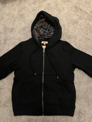 Burberry hoodie full zip - Snygg svart hoodie från Burberry med hel dragkedja och klassiskt rutigt foder i huvan och insidan. Hoodien har två stora fickor framtill och justerbar dragsko i huvan. Materialet är mjukt och bekvämt, perfekt för en avslappnad stil. Helt ny inte använt eller nåt