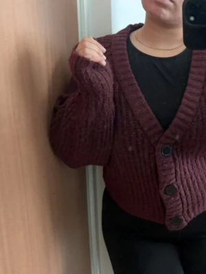 Vinröd chunky kofta med stora knappar - Mysig vinröd kofta i chunky stickat material med bred ribbstickning och stora svarta knappar framtill. Koftan har en djup V-ringning och långa ärmar med avslappnad passform. Perfekt för lager på lager och en chill look.