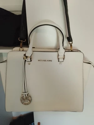 Vit handväska från Michael Kors NYSKICK - Snygg vit handväska från Michael Kors i stilren design med svarta detaljer och guldfärgade metalldelar. Väskan har både handtag och en avtagbar axelrem samt MK-logga som hänge. Tillverkad i slätt konstläder och passar perfekt till en trendig look. NYSKICK