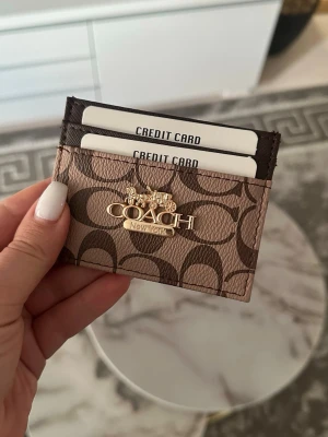 Coach brun korthållare i läder - Snygg korthållare från Coach i brunt läder med klassiskt monogrammönster och guldfärgad logga framtill. Perfekt för att organisera dina kort på ett stilrent sätt. Smidig och tunn design som enkelt får plats i fickan eller väskan.