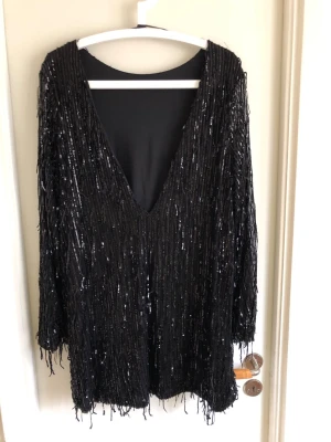 Svart glittrig paljettklänning - Snygg svart klänning med djup v-ringning och långa ärmar. Klänningen är täckt av glittriga paljetter som ger en festlig look. Perfekt för dig som vill sticka ut och glänsa. Materialet känns mjukt mot huden och modellen är kort.