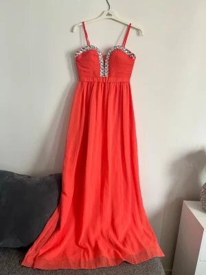 Korall långklänning med strass - Snygg korall långklänning med tunna axelband och djup v-ringning dekorerad med stora strassdetaljer. Klänningen har markerad midja och ett flytande, lätt tyg som ger en elegant siluett. Perfekt för speciella tillfällen.