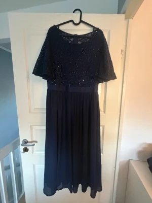 Mörkblå långklänning med spets och glitter - Elegant mörkblå långklänning med glittrig spets upptill och fladdrande korta ärmar. Kjolen är i ett lätt, fallande tyg och midjan markeras av ett satinband. Perfekt för speciella tillfällen och bal.