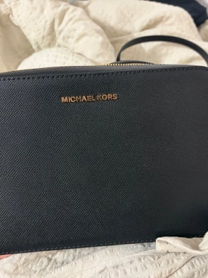 Svart axelväska Michael Kors - Snygg svart axelväska från Michael Kors i strukturerat läder med guldfärgade detaljer och kedja på axelremmen. Väskan har en stilren och enkel design med dragkedja upptill och logga i guld framtill. Perfekt för dig som gillar klassisk och trendig stil. Väskan är helt oanvänd, fick den sen present och där av inte kunde lämna tillbaka den.