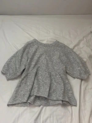 Grå peplumstickad tröja från Nelly - Supersöt grå stickad tröja från Nelly i peplummodell. Tröjan har rund halsringning, trekvartsärmar och en volang nedtill som ger en snygg siluett. Perfekt att styla med jeans eller kjol för en trendig look.