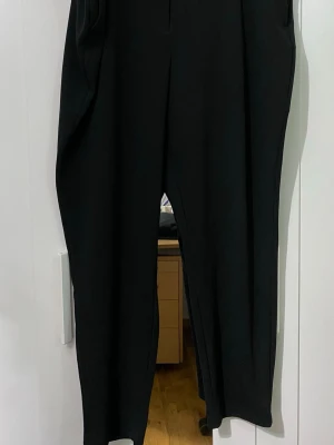 Svarta kostymbyxor med rak passform - Snygga svarta kostymbyxor med rak passform och pressveck. Byxorna har hög midja och sidfickor. Perfekta för en stilren look. Materialet ser ut att vara en mjuk och följsam textil, troligtvis polyesterblandning. Som ni ser står det ingen storlek men jag gissar på att den är XXL